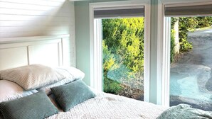Un dormitorio con cama, almohadas y vistas a la vegetación a través de amplias ventanas.