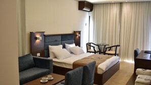 A hotel room with a bed, two armchairs, a small table with cups, a desk with a chair, and a window with curtains. - Sands Hotel (Byblos)