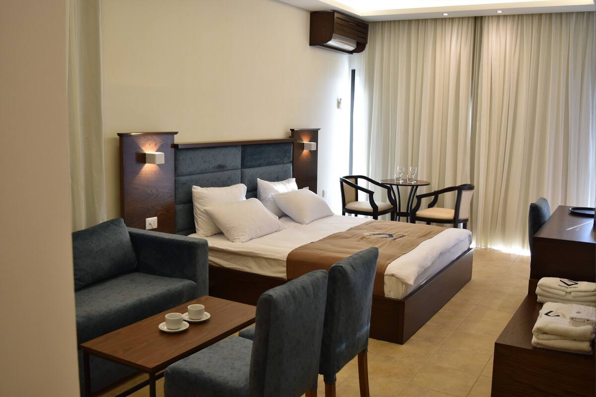 A hotel room with a bed, two armchairs, a small table with cups, a desk with a chair, and a window with curtains.