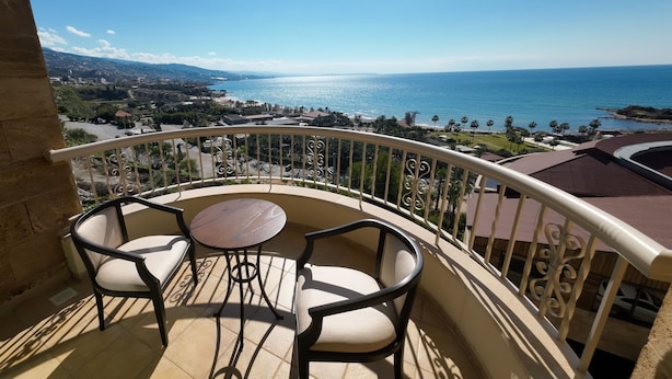 A balcony with two chairs and a small table, overlooking a coastal landscape with a view of the sea and distant hills. - Sands Hotel (Byblos)