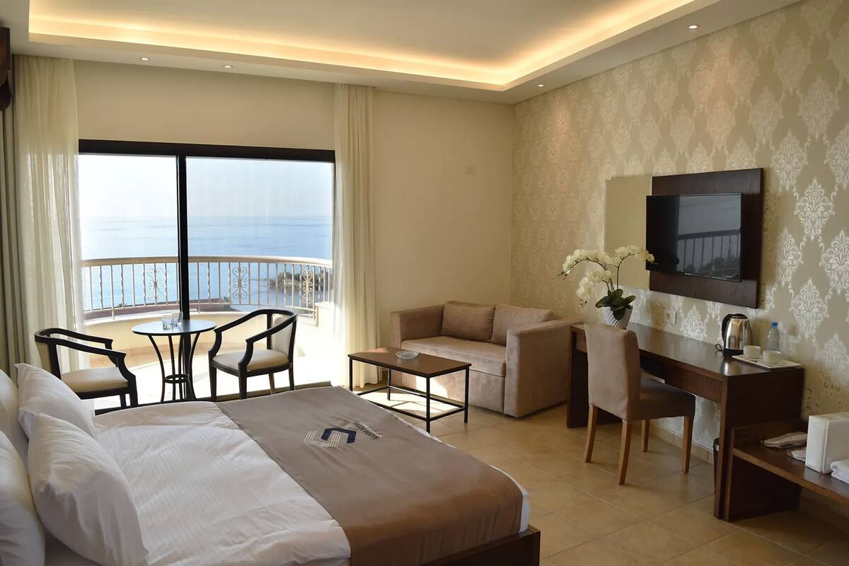 A hotel room with a bed, a desk, a sofa, a TV, and a balcony with a view of the sea.