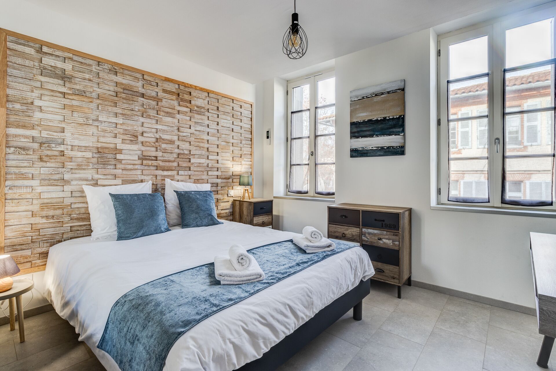 Quarto moderno com parede de madeira, cama com roupa de cama branca e azul, mesa de cabeceira com abajur e vista para edifícios através de janelas amplas.