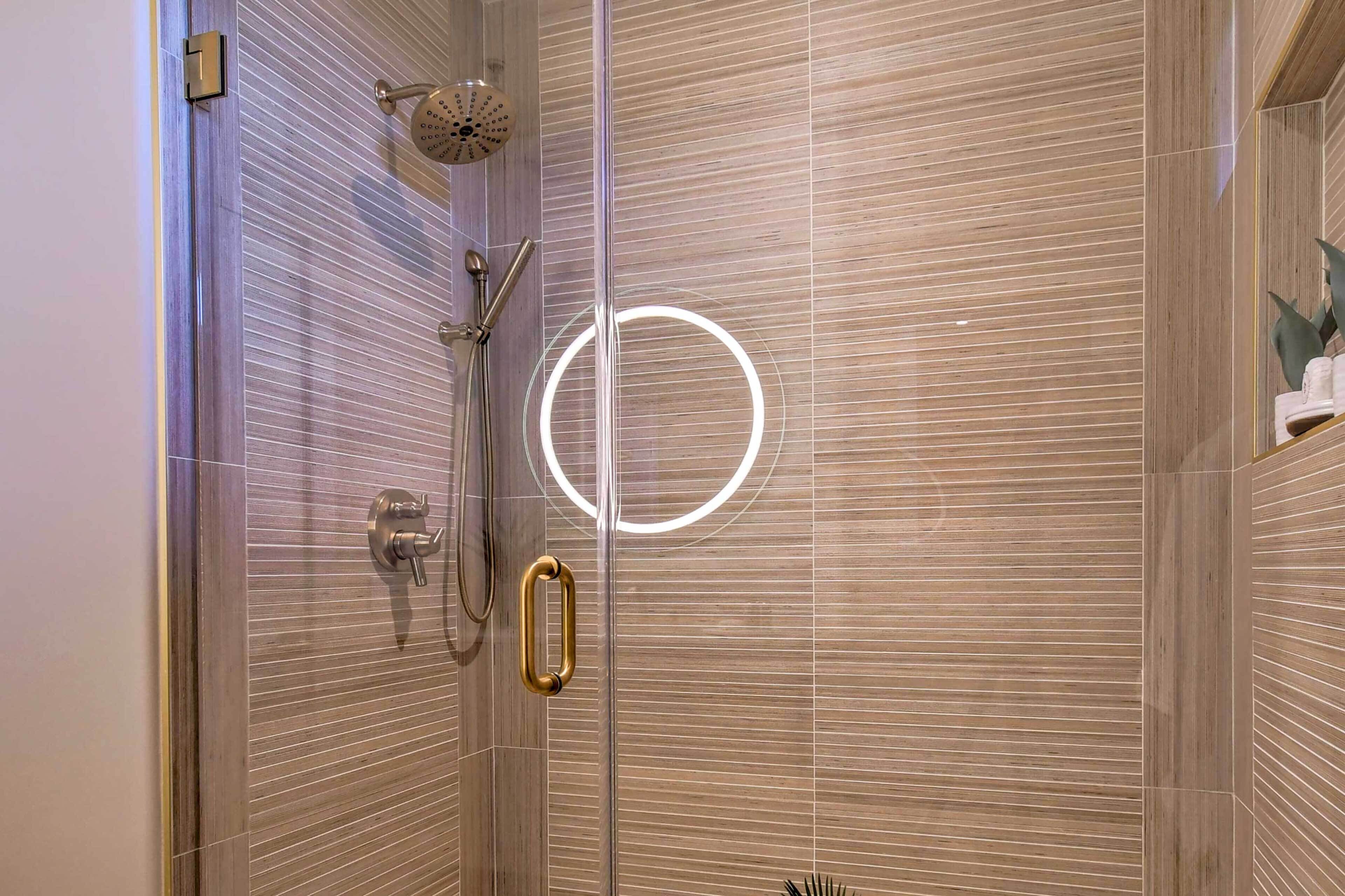 Een moderne douche met een glazen deur, een gouden hendel en een ronde lamp op de muur.