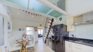 Una cocina moderna con un refrigerador grande, un comedor con mesa de madera y sillas blancas, y una escalera que sube a un nivel superior.