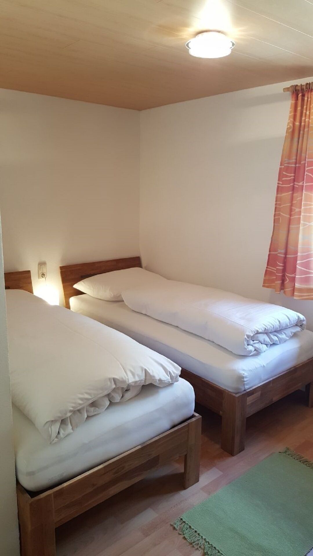 Una habitación con dos camas de madera, un tapete verde y una ventana con cortinas naranjas.