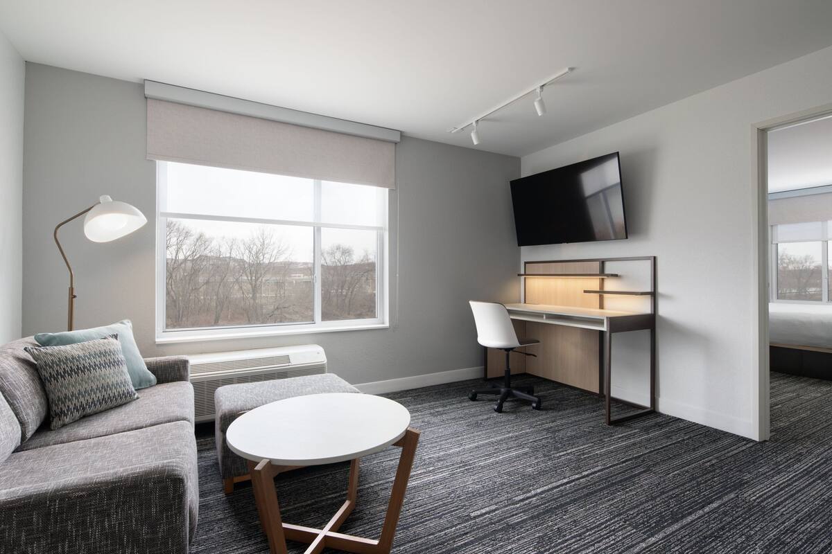 Une pièce moderne avec un canapé gris, une table ronde, un bureau avec une chaise, un lampadaire et un téléviseur fixé au mur.