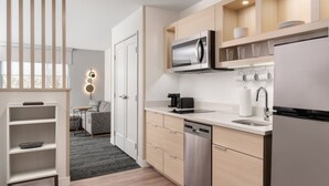 A modern kitchen with a built-in microwave, dishwasher, and a coffee maker. There is a wooden shelf above the sink and a small seating area with a sofa and table in the background. - Towneplace Suites By Marriott New Philadelphia (New Philadelphia)