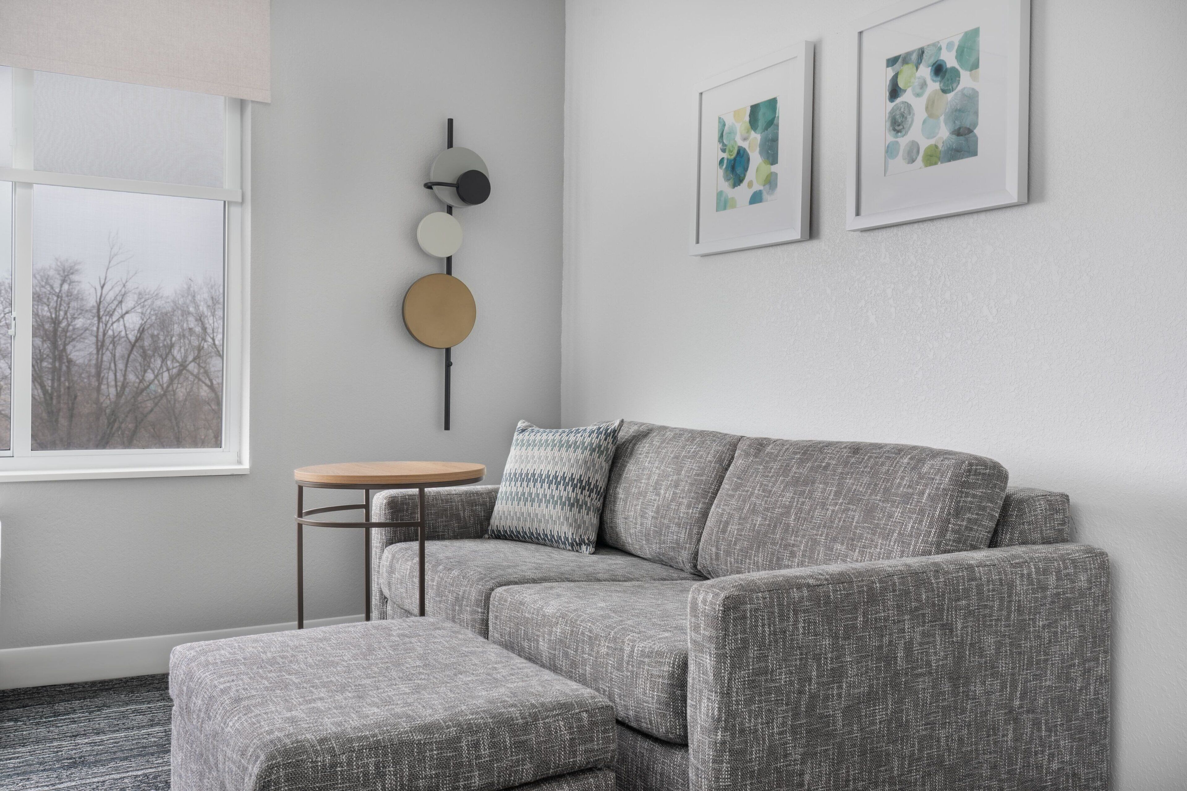 A modern living room with a grey sofa, a round wooden table, and a grey patterned ottoman. There are abstract paintings on the wall and a wall-mounted light fixture.
