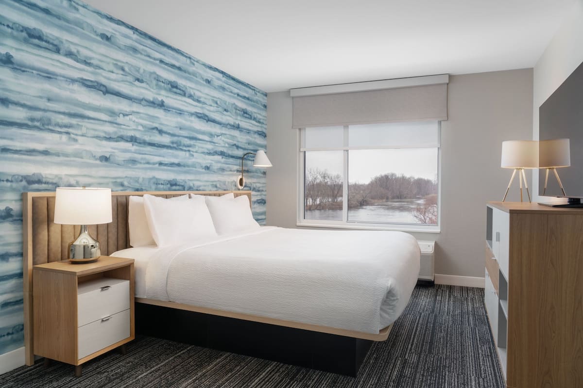 Une chambre d’hôtel moderne avec un grand lit, une table de chevet avec une lampe, une commode et une fenêtre donnant sur des arbres.