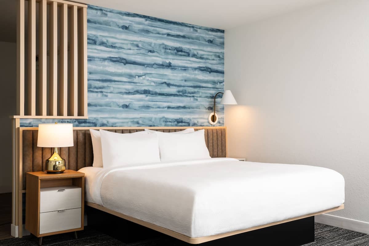 Une chambre d’hôtel avec un lit, une table de chevet avec une lampe, un mur orné d’un motif abstrait bleu et un luminaire fixé au mur.