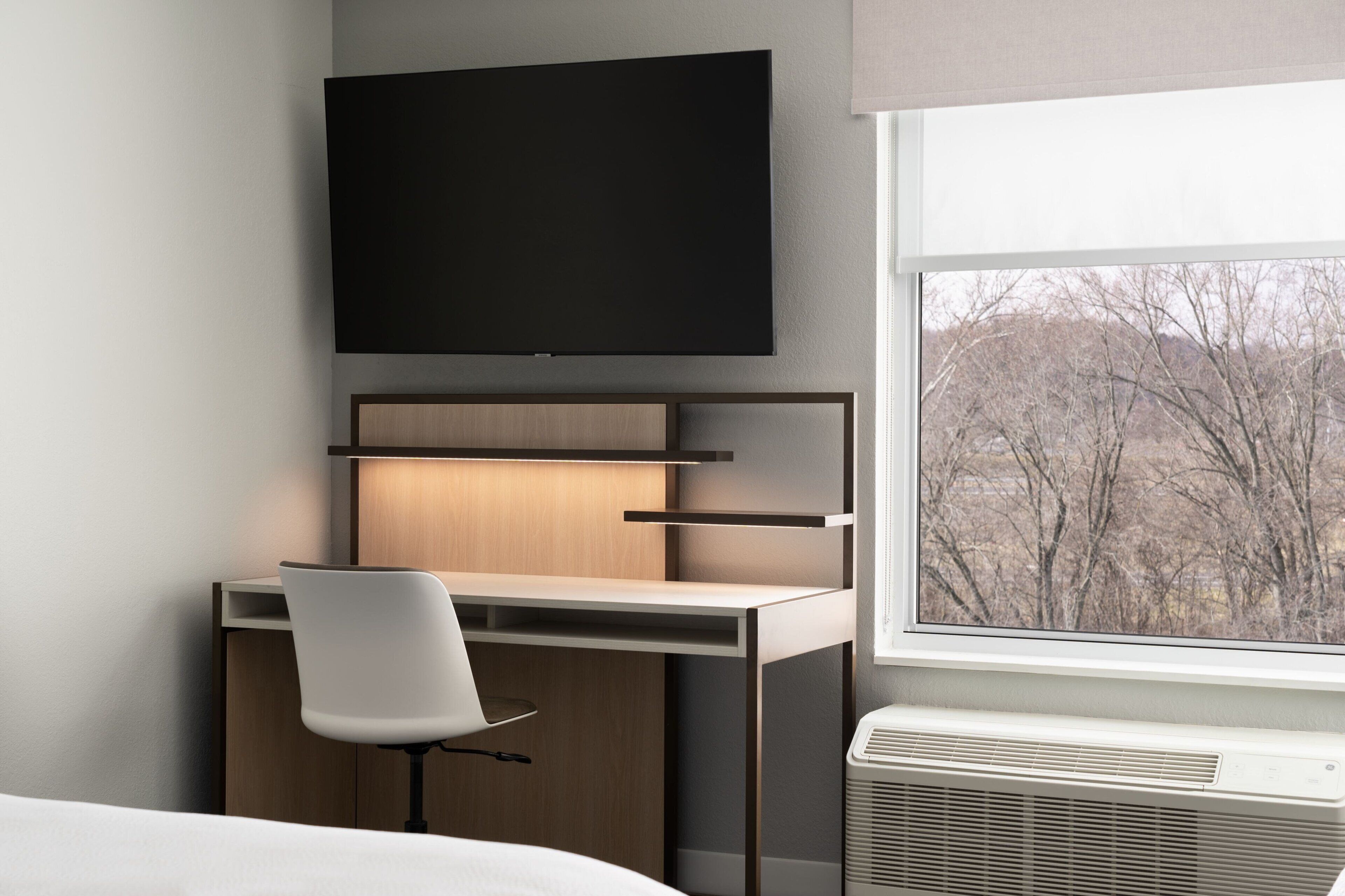 A modern hotel room with a desk, chair, flat-screen TV, and a window with a view of trees.