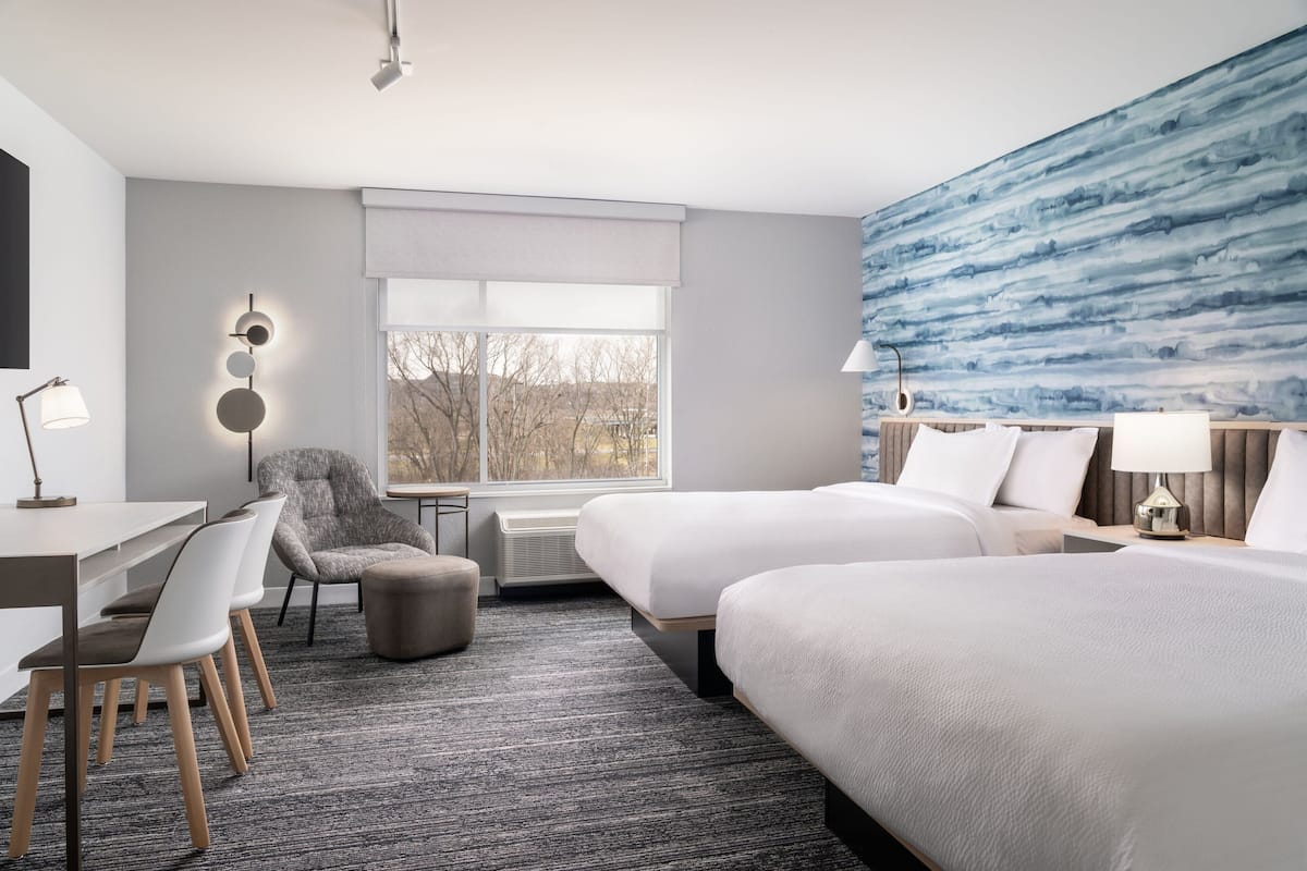 Une chambre d’hôtel avec deux lits, un bureau, une chaise, une lampe et une grande fenêtre.