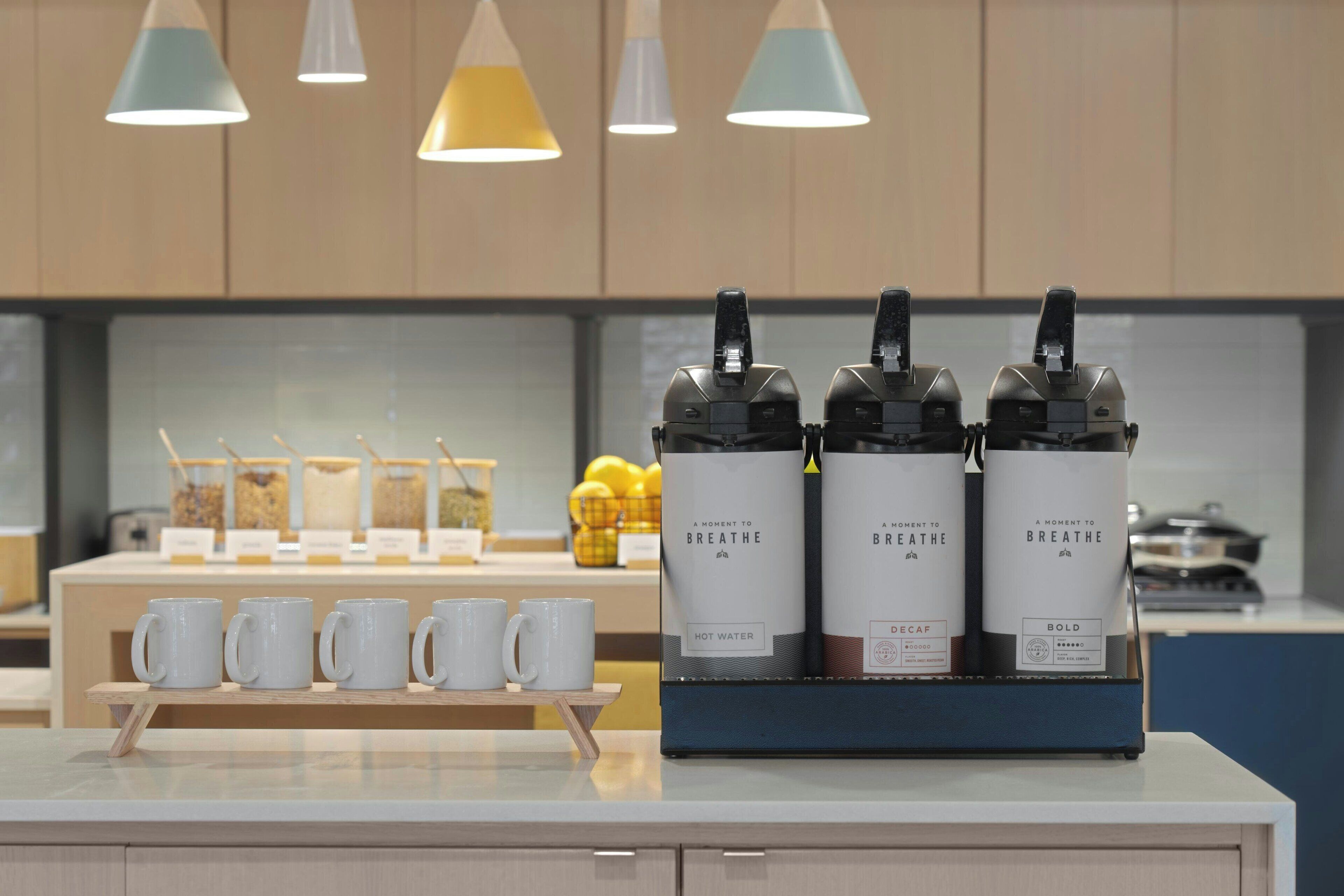 A coffee station with three coffee dispensers labeled "A Moment to Breathe" and a selection of white mugs on a wooden tray.