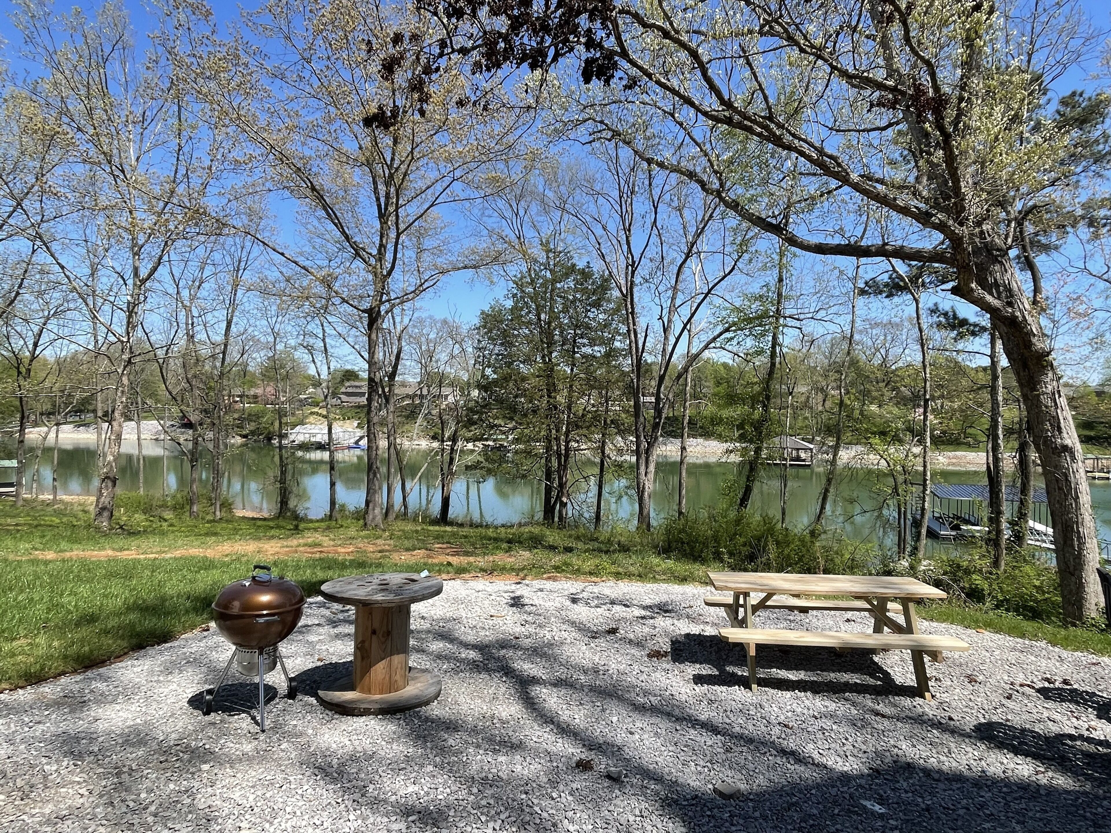 Een picknickplaats met een barbecue, een tafel en bankjes, nabij een meer omringd door bomen.