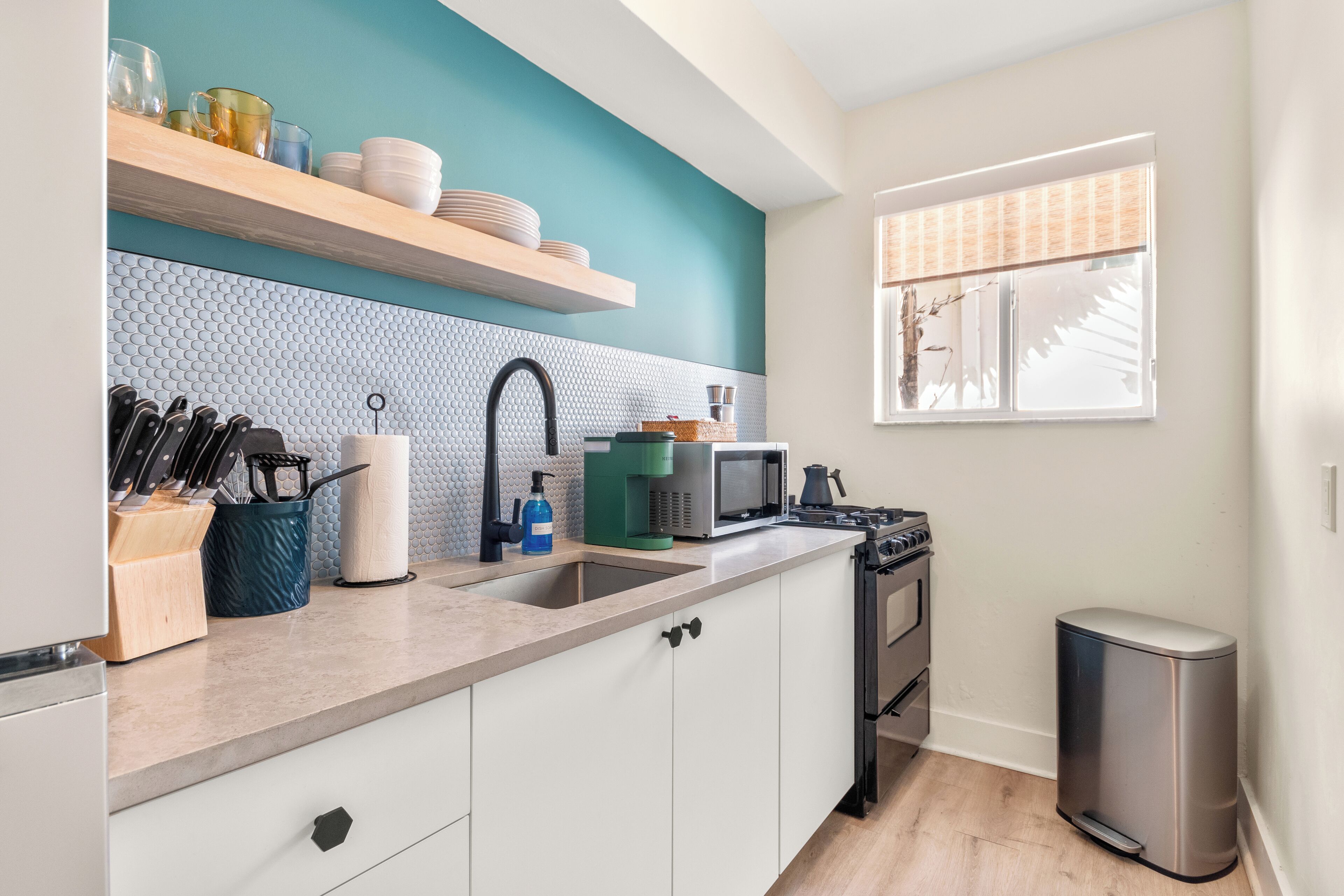 A modern kitchen with white cabinets, a teal accent wall, a stainless steel sink, and a window with blinds.