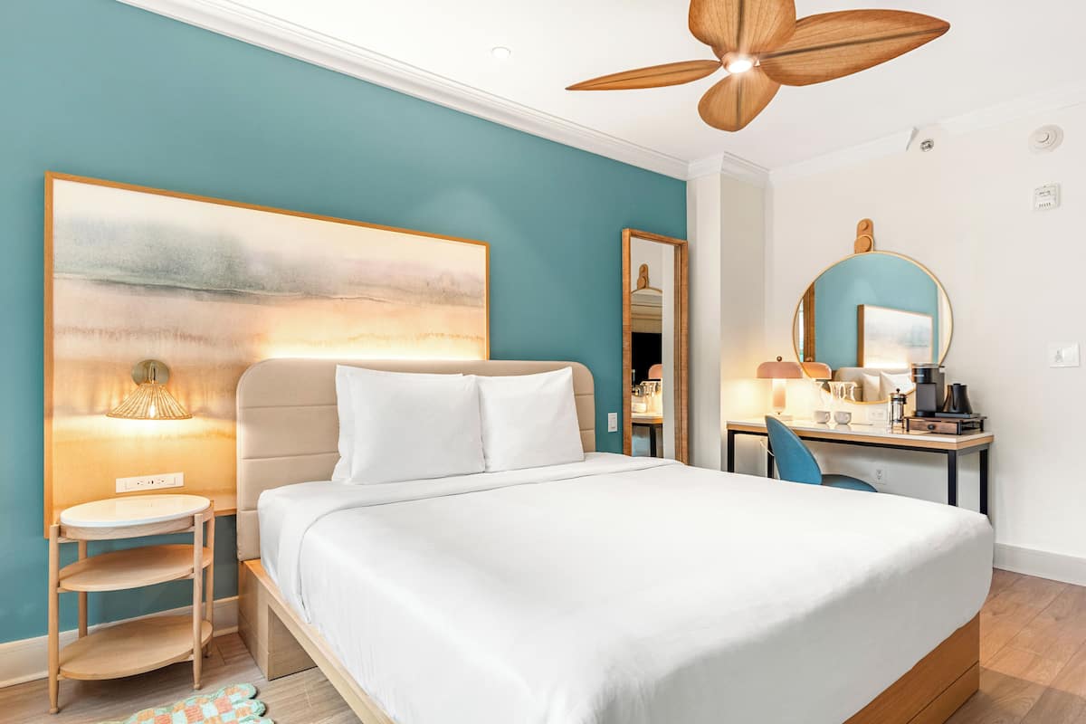 Une chambre à coucher comprenant un lit, un bureau, une chaise, un ventilateur de plafond, un miroir fixé au mur et un tableau.