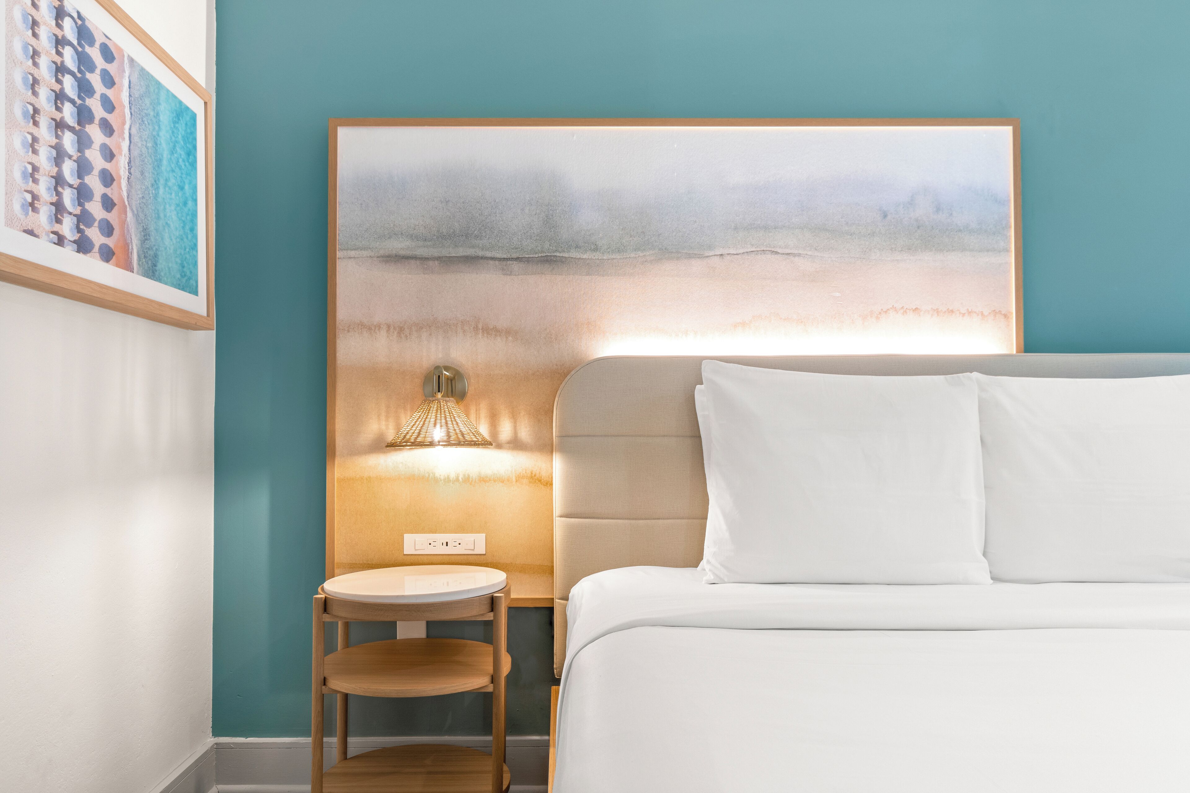 Quarto de hotel com cama, mesa de cabeceira, abajur e um quadro na parede.