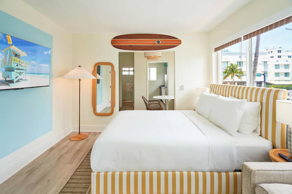 Une chambre d’hôtel avec un grand lit, un coin salon avec des coussins à rayures, un téléviseur à écran plat, une lampe sur pied et une vue sur la ville par la fenêtre.
