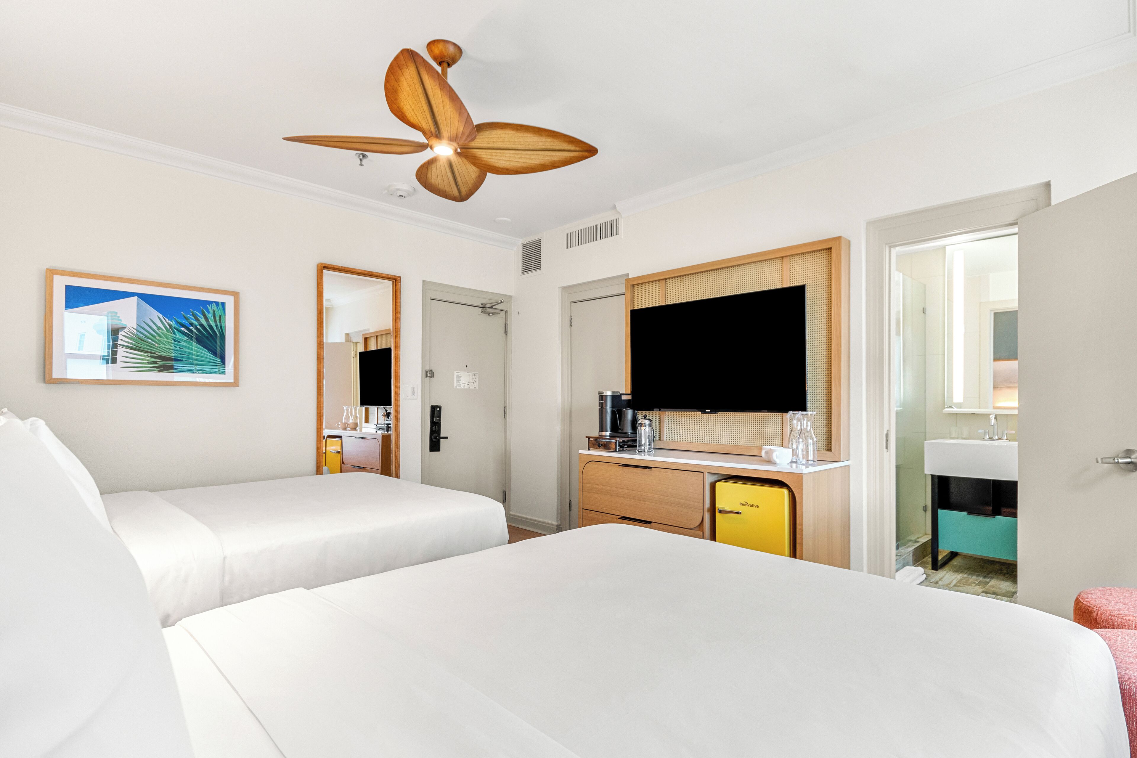 Quarto de hotel com duas camas, um ventilador de teto de madeira, um espelho grande, uma TV de tela plana, um minibar e um banheiro visível por uma porta aberta.