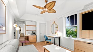 Una habitación moderna y bien iluminada con ventilador de techo, sofá, mesa de comedor, escritorio con silla, espejo y televisor.