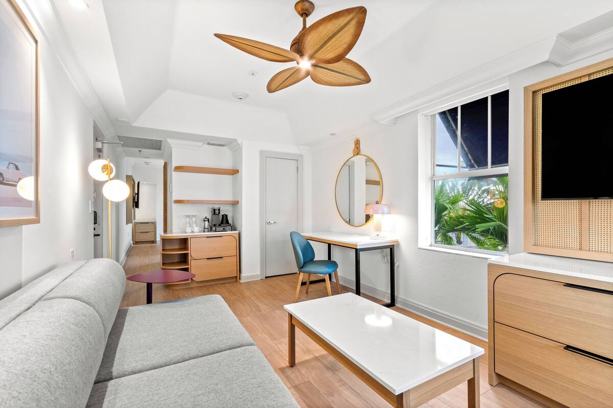 Une pièce moderne et bien éclairée, dotée d’un ventilateur de plafond, d’un canapé, d’une table à manger, d’un bureau avec une chaise, d’un miroir et d’un téléviseur.