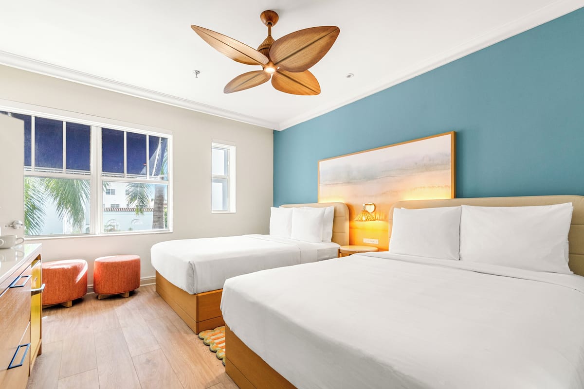 Une chambre d’hôtel avec deux lits, un ventilateur de plafond, une grande fenêtre donnant sur des palmiers et un tableau au mur.