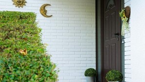 Une façade en briques blanches avec une porte sombre, un décor en forme de croissant de lune et deux plantes en pot placées sur les marches.