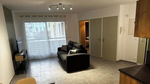 A modern living room with a sofa, a TV, and a dining area. There is a large window with blinds, a door leading to a bedroom, and a kitchen area with cabinets.