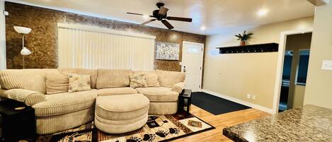 A living room with a sectional sofa, ottomans, a ceiling fan, and a wooden floor.