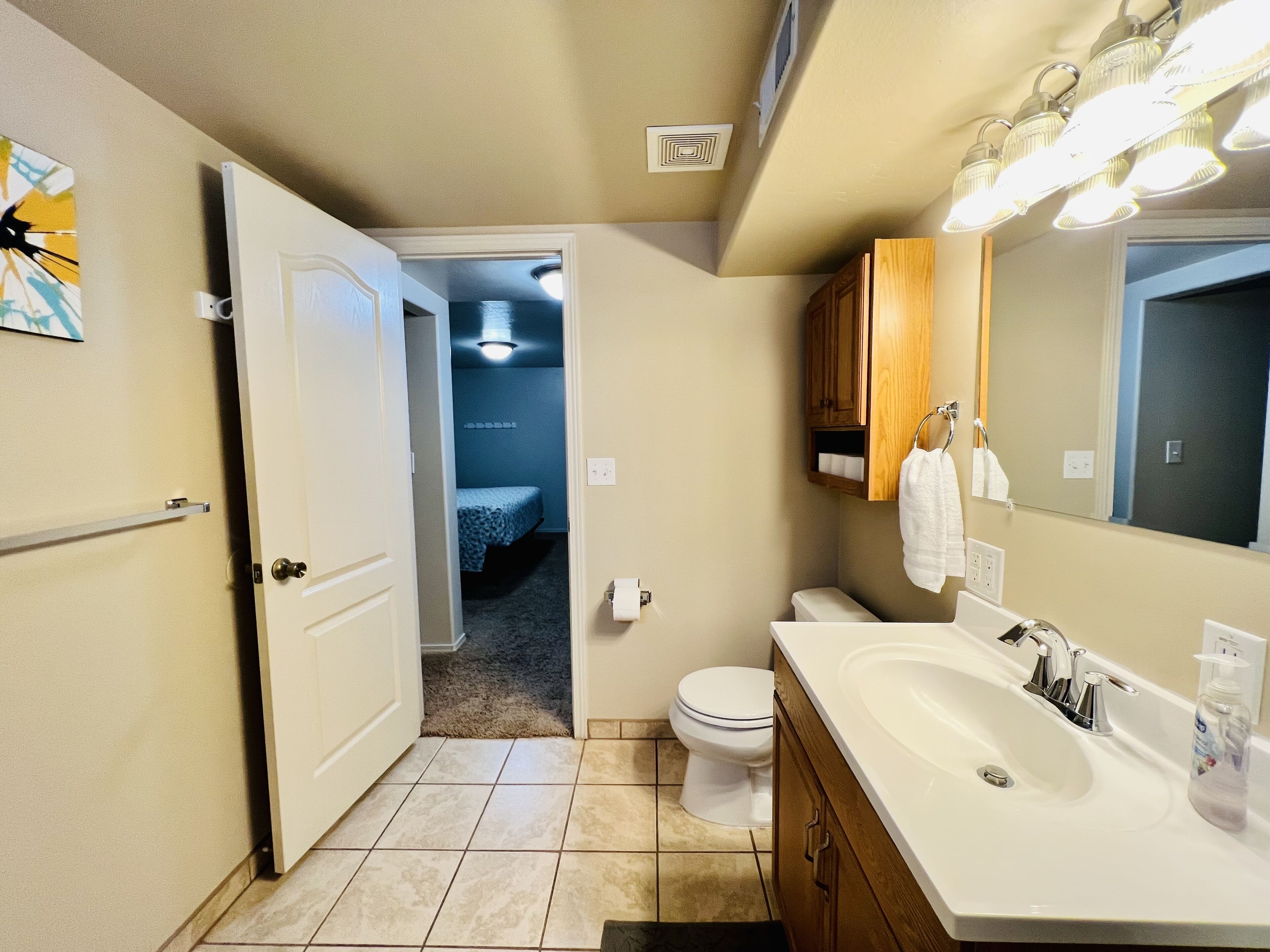 A bathroom with a white sink, mirror, and toilet. There is a towel rack and a small wooden cabinet above the toilet. The floor is tiled, and there is a door leading to another room.
