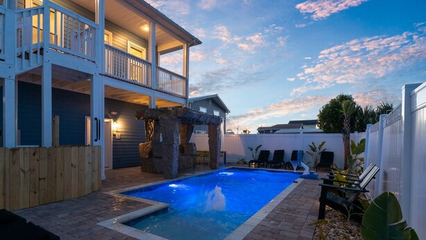 Una casa de dos pisos con piscina, terraza de madera y muro de piedra.