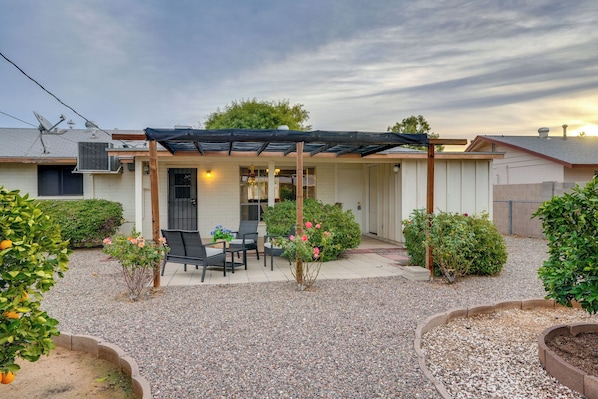 Relax in the serene backyard with a covered patio, table, and chairs, surrounded by lush greenery and a small tree with vibrant flowers.