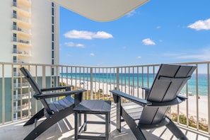 Ein Balkon mit zwei Stühlen und einem Tisch, mit Blick auf einen Strand und klares, blaues Wasser sowie einige Wolken am Himmel.