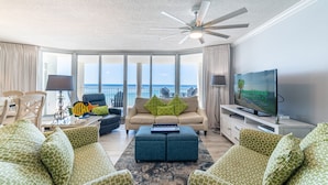 A spacious living room with a large window overlooking the ocean, a ceiling fan, and a flat-screen TV on a white stand. - Edie's Escape-Long Beach 501T3-3BD/3BTH End Unit-2 Sets of Beach Chairs Included! (Panama City Beach)