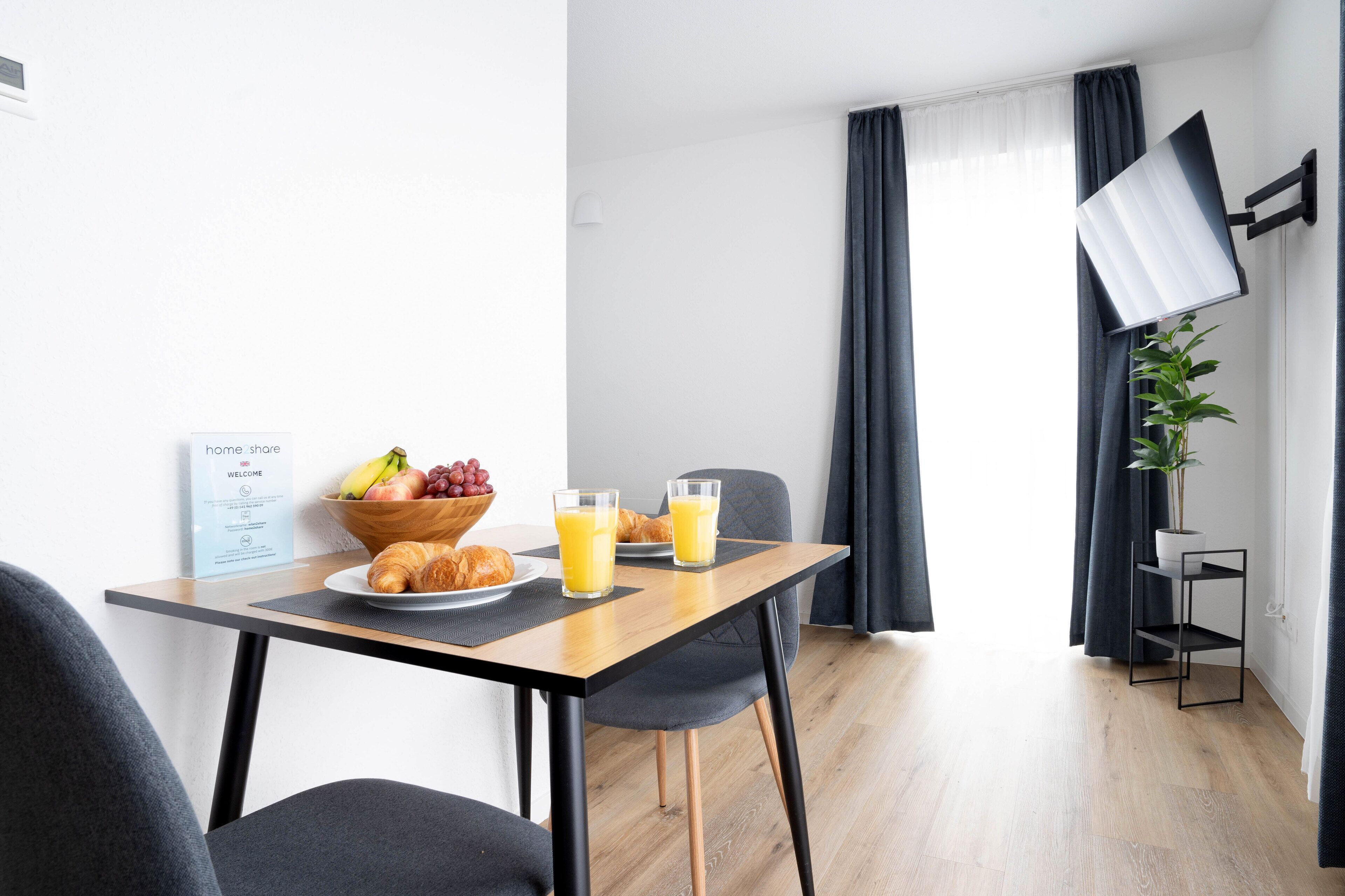 Una habitación de hotel moderna con una mesa de comedor de madera, dos sillas, un cuenco de fruta y dos vasos de jugo de naranja.