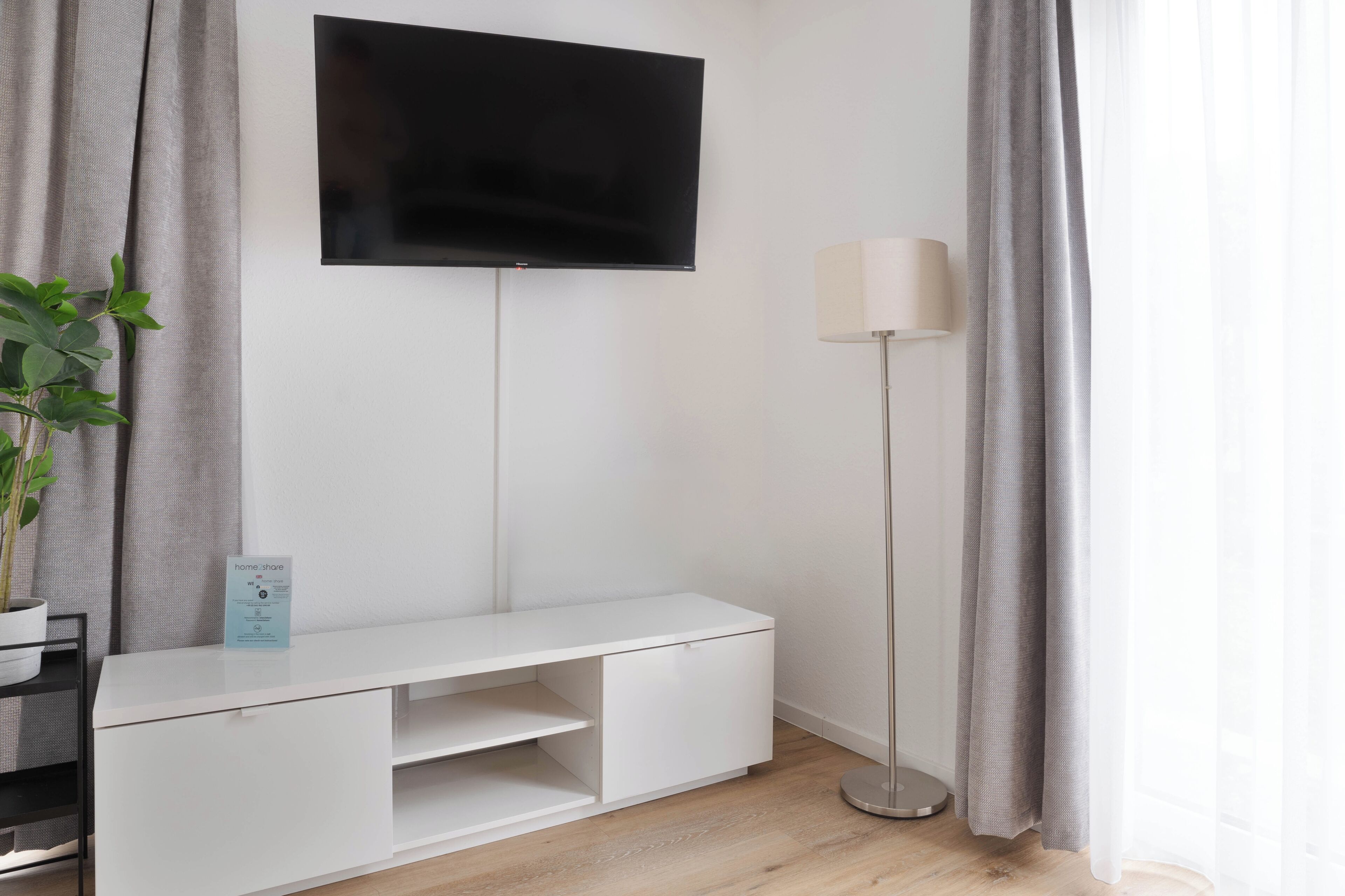 A modern living room with a flat-screen TV mounted on the wall, a white entertainment unit, a floor lamp, and a potted plant.