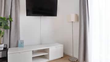 A modern living room with a flat-screen TV mounted on the wall, a white entertainment unit, a floor lamp, and a potted plant.