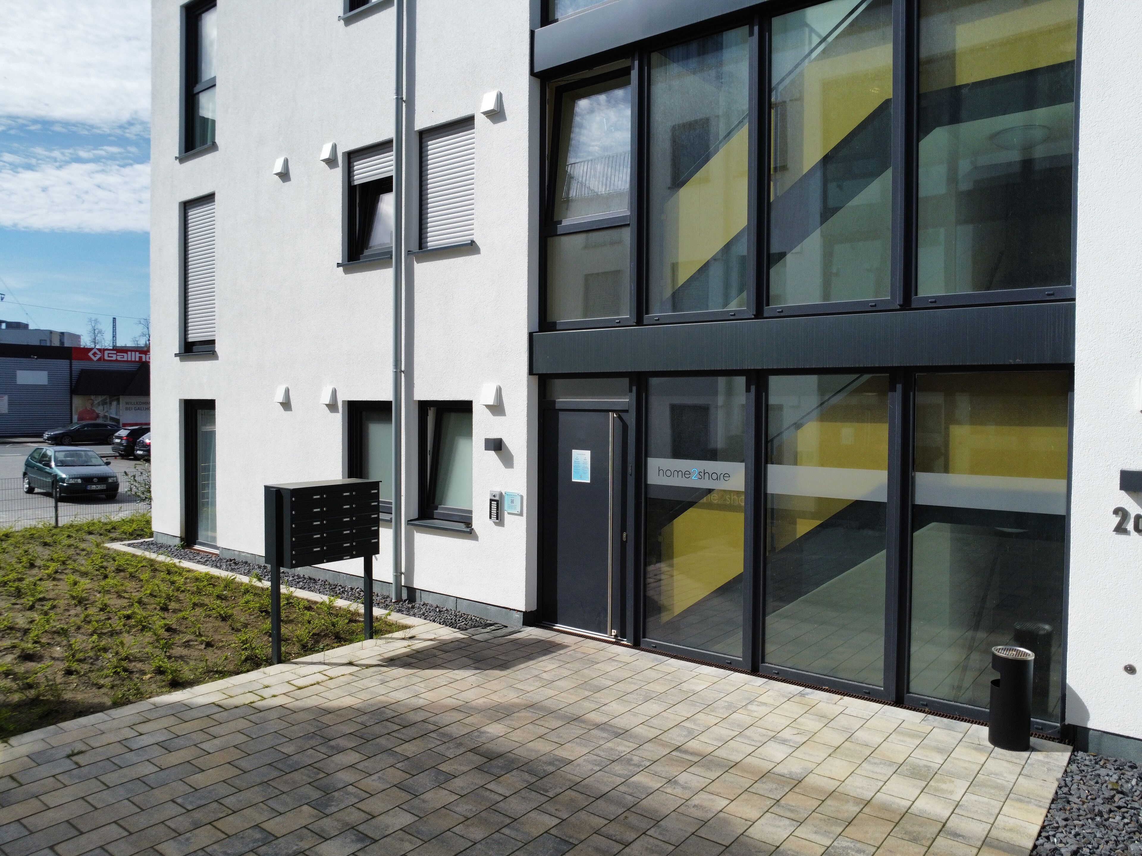 Un bâtiment moderne avec une façade en verre, une porte noire portant l’inscription « home2share », et un petit espace extérieur comprenant une boîte aux lettres et une poubelle.
