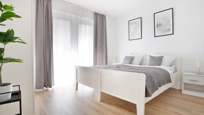 A modern bedroom with a white bed, grey curtains, a bedside table, and a potted plant. - Deluxe apartment in the centre of OS (Osnabrueck)