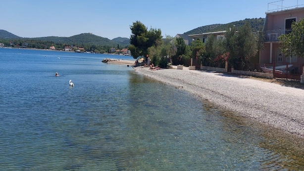 A coastal area with a pebbly beach, clear blue water, and a row of buildings along the shore. - Apartment by the Beach (Bilice)