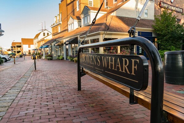 Un banco con la inscripción 'BOWEN'S WHARF', situado en una calle empedrada de ladrillo y edificios al fondo.