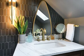Un baño con lavamanos de mármol blanco, grifería dorada, una planta en maceta y un salpicadero de azulejos hexagonales.