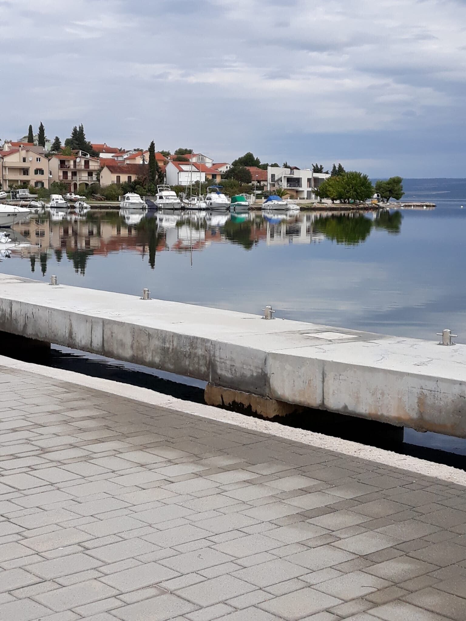 Uma marina com barcos atracados, um calçadão de concreto e edifícios ao fundo.