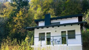 Un edificio blanco de dos plantas con techo oscuro, rodeado de abundante vegetación.