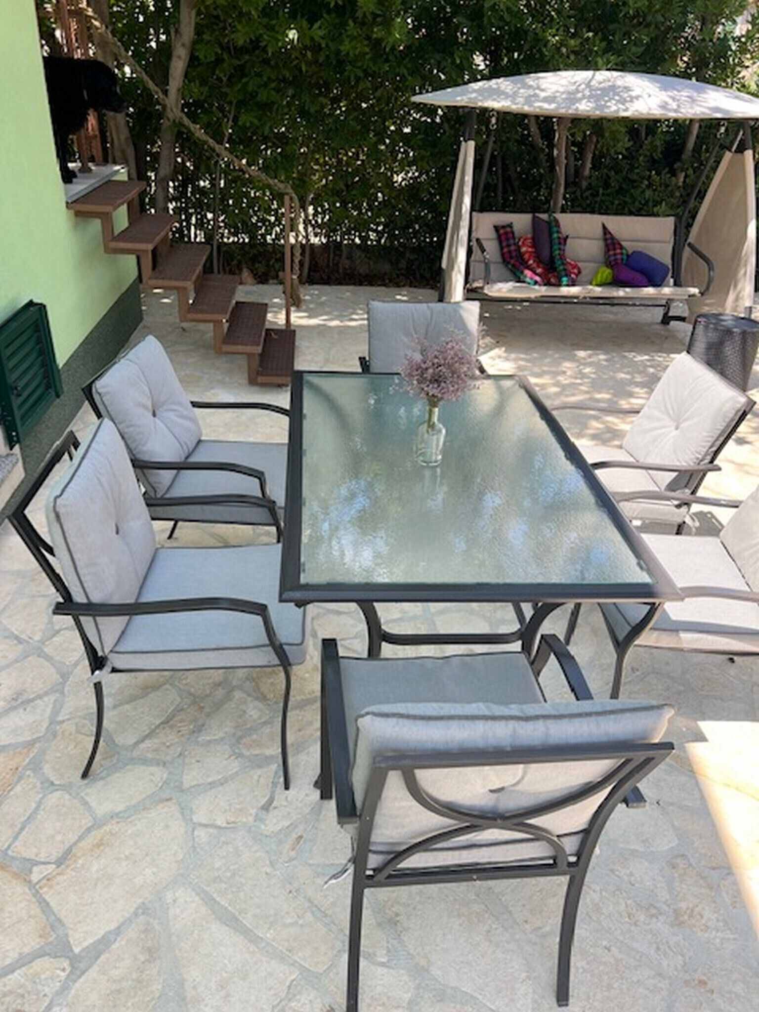 An outdoor patio with a glass-top table surrounded by cushioned chairs, a small vase with flowers, and a swing set in the background.