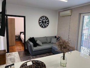 Uma sala de estar moderna com um sofá cinzento, um relógio de parede redondo e um vaso com flores secas sobre uma mesa branca.