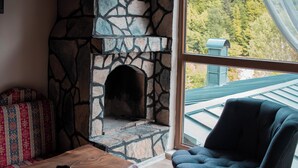 Una chimenea de piedra, una mesa de centro de madera, un sofá estampado y un sillón oscuro en una habitación con vistas a un paisaje verde a través de una ventana.