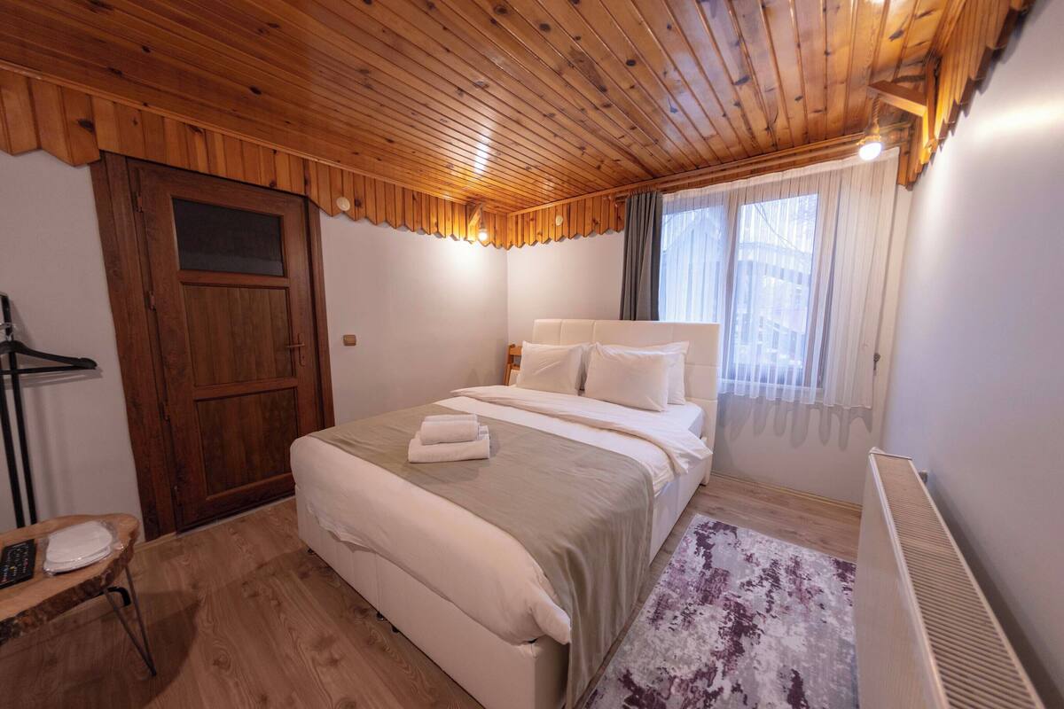 A bedroom with a wooden ceiling, a bed with white bedding, a radiator, and a window with curtains.