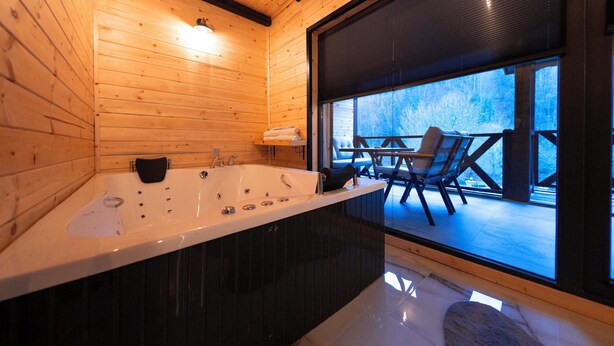 Interior de una cabaña de madera con una bañera grande, una zona de estar con sillones y vistas al exterior a través de un ventanal amplio.
