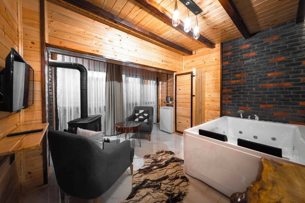 A modern log cabin interior with a large wooden ceiling, a black leather sofa, a round wooden table, a white bathtub, and a brick accent wall.